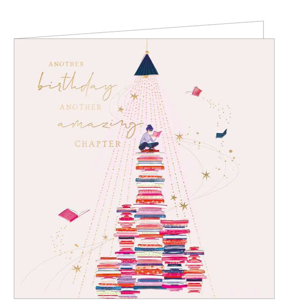 This light-pink birthday card is decorated with an illustration of a young woman sitting atop several piles of books as more books and stars fly around her. Gold text on the front of the card reads 