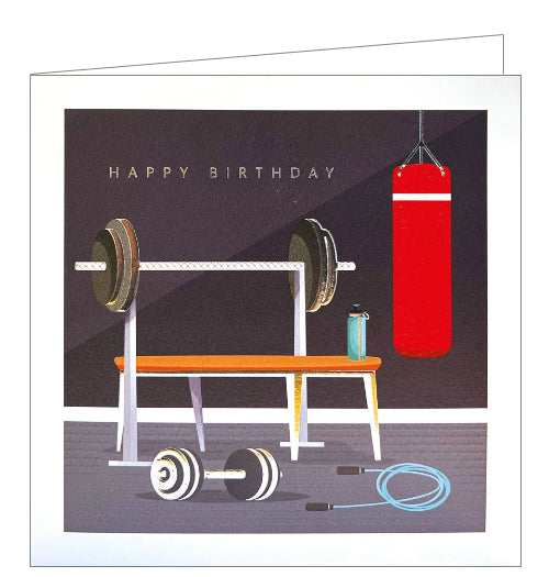 This birthday card is the perfect choice for a fitness fan! This card is decorated with a bench and barbell on a rack, a punching bag hanging from the ceiling, and a dumbell and skipping rope on the floor. Silver text on the front of the card reads 