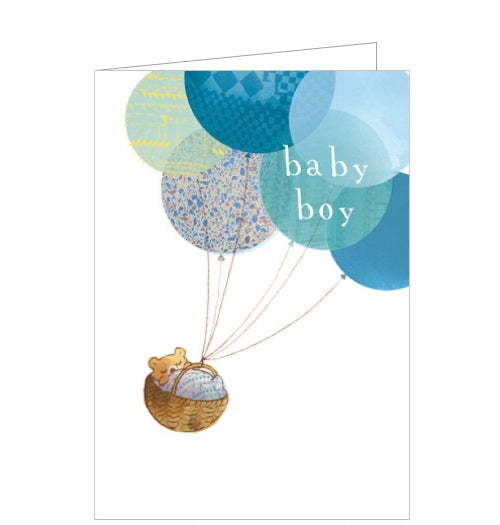 This sweet new baby boy card is decorated with an illustration of a baby bear cub asleep in a basket tied to a bunch of blue balloons,  White text on the balloons reads 