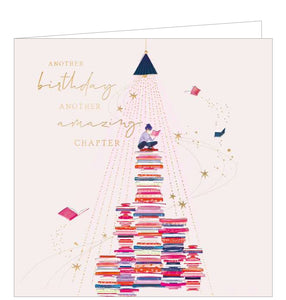 This light-pink birthday card is decorated with an illustration of a young woman sitting atop several piles of books as more books and stars fly around her. Gold text on the front of the card reads "Another Birthday, Another amazing chapter".