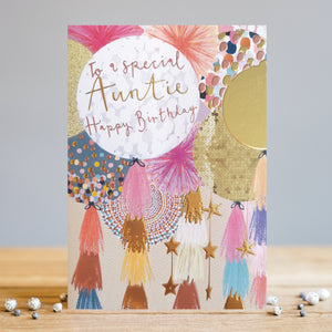 This birthday card for a special aunty features a modern and eye-catching design with gold detailing. This birthday card is decorated with a close-up illustration of colourful balloons in shades of pink, peach and gold. Each balloon has tassels and stars hanging from their strings.The text on the closest balloon reads "To a Special Auntie Happy birthday".
