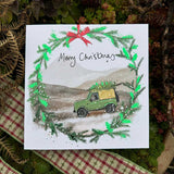 Christmas card by artist Alex Clark decorated with a christmas wreath. Within the wreath a back dog appears to drive a 4 x 4 car through a snowy moor - with a Christmas tree tied to the roof of the car. The text on the front of the card reads "Merry Christmas x".