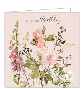 This is a stunning birthday card decorated with an illustration of a bumble gathering nectar from dreamy pink and gold flowers. Gold text in the top corner of the card reads "On your Birthday".