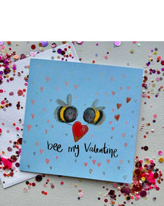 A wonderfully cute Valentine's card by Alex Clark, featuring two romantic bees holding red heart between them. The background is blue and dotted with mini heart details. Black script on the front of the card reads "bee my Valentine".