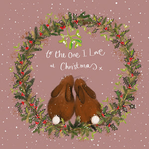 Part of Alex Clark's Christmas card collection, this Christmas card is decorated with a festive wreath, inside of which two rabbits snuggle together under the mistletoe. White text on the front of the card reads "to the One I Love at Christmas x".