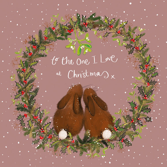 Part of Alex Clark's Christmas card collection, this Christmas card is decorated with a festive wreath, inside of which two rabbits snuggle together under the mistletoe. White text on the front of the card reads 