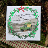 Christmas card by artist Alex Clark decorated with a christmas wreath. Within the wreath if a scene of a black labrador dog seemingly driving a landrover - with a christmas tree on its roof - across a snowy moor. THe text on the front of the card reads "Merry Christmas to a Wonderful Grandson x" in a hand-written style font.