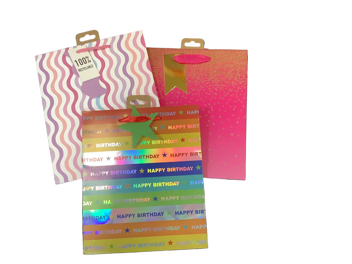 Three colorful birthday-themed medium sized gift bags. One has holographic rainbow stripes with 'HAPPY BIRTHDAY' text, one is pink with gold speckles, and one has pink, purple, and white wavy stripes with a small cup graphic.