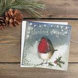 Part of Alex Clark's Christmas card collection, this petite Christmas card is decorated with Alex's painting of a red-cheeked, red-breasted fluffy robin perched on a branch of holly. White text on the front of the card reads "Thinking of you".