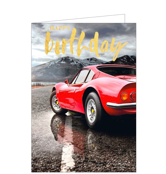 This birthday card is decorated with a photograph of a red sports car stopped on a mountain road. Gold text on the card reads 