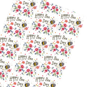 This Alex Clark wrapping paper features a repeating design of a smiling bumble bee carrying a flower, surrounded by brightly coloured flowers and text that reads "Happy Bee Day". 