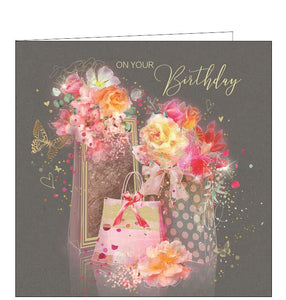 This birthday card is decorated with pink and gold gift bags overflowing with flowers. Metallic gold text on the front of the card reads "On your Birthday".