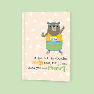 Ideal for keeping notes on anything from workouts to recipes, this this softbacked A6 lined notebook is decorated with a brown bear wearing sweatbands and sports kit and holding cookies. The caption on the front of the note book reads "if you eat the cookies really fast, fitbit may think you are running...". 