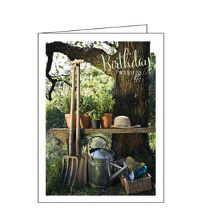 This birthday card is decorated with a photograph of a garden bench, beside a tree, laiden with potted plants and garden tools. The text on the front of the card reads "Birthday Wishes".