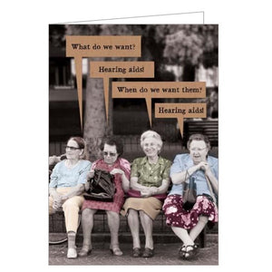 This funny blank card features a vintage colourized photograph of four older women sitting on a park bank.. Four speech bubbles on the card read "What do we want?" "Hearing aids!" "When do we want them?" "Hearing aids!"