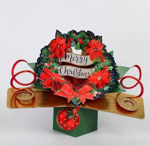 This pop-up 3D keepsake Christmas card is decorated with a 3d Christmas wreath of red poinsettia flowers and green foliage. A banner at the centre of the wreath reads "Merry Christmas".